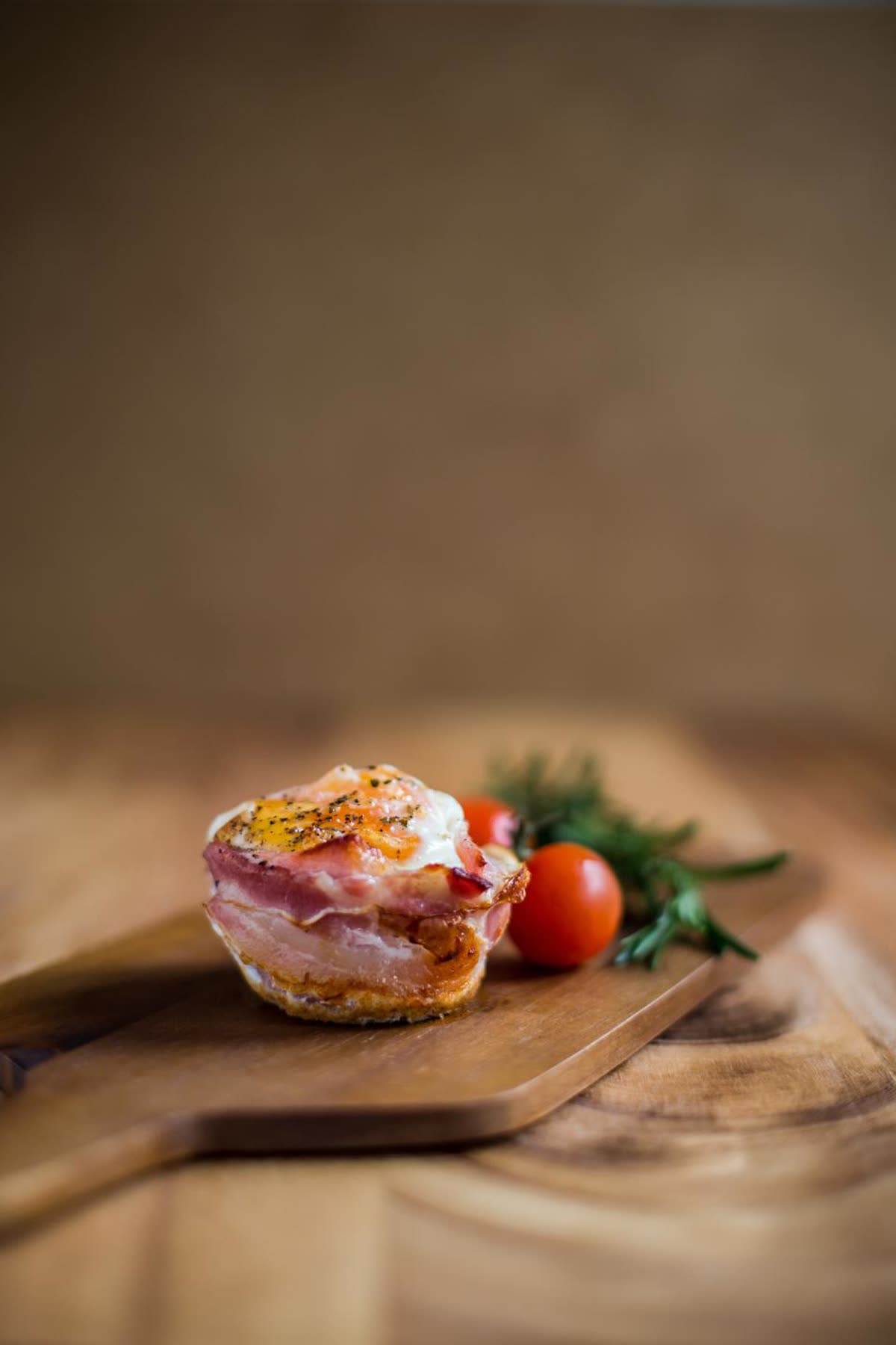 A bacon-wrapped baked egg on a wooden board, with tomatoes behind